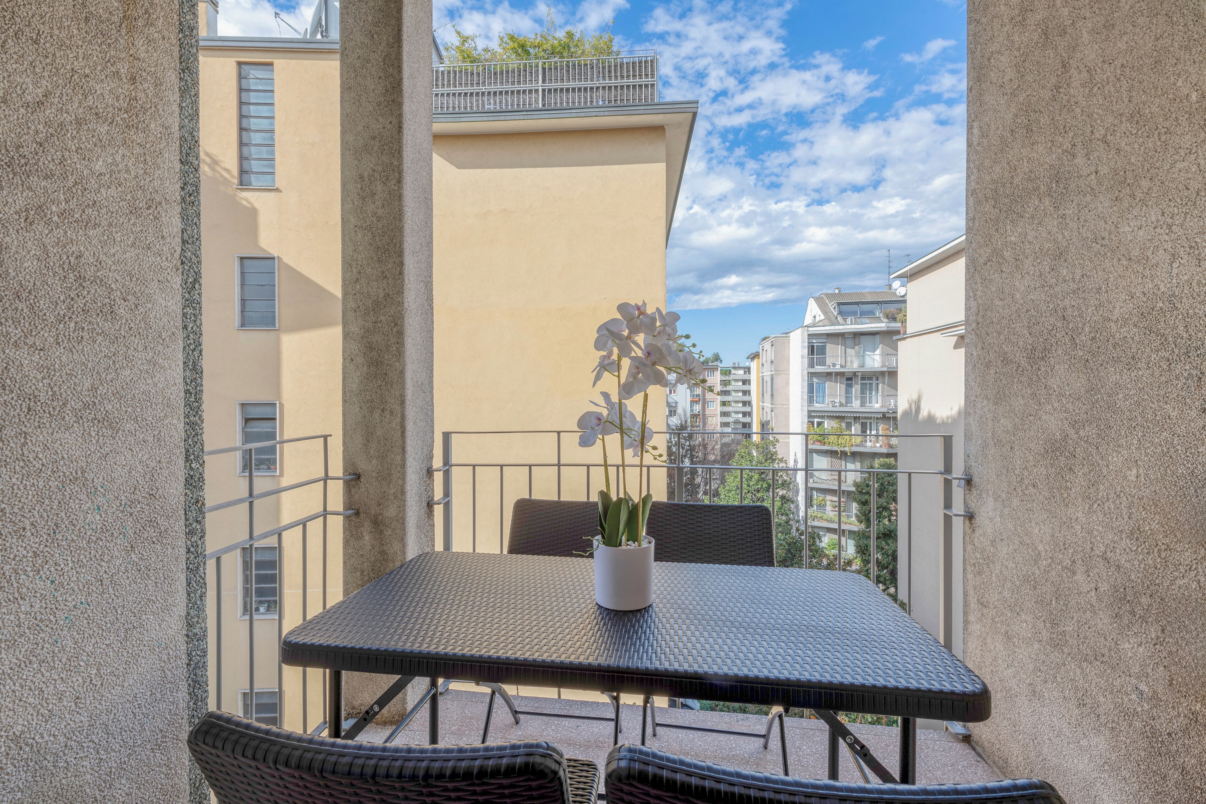 En balkon med et bord, stole, en potteplante og udsigt til omkringliggende bygninger.