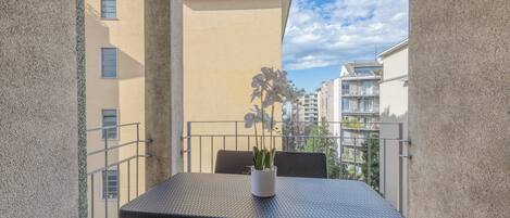 En balkon med et bord, stole, en potteplante og udsigt til omkringliggende bygninger.