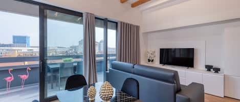 A modern living room with a glass dining table, black chairs, a blue sofa, and a TV on a white cabinet.