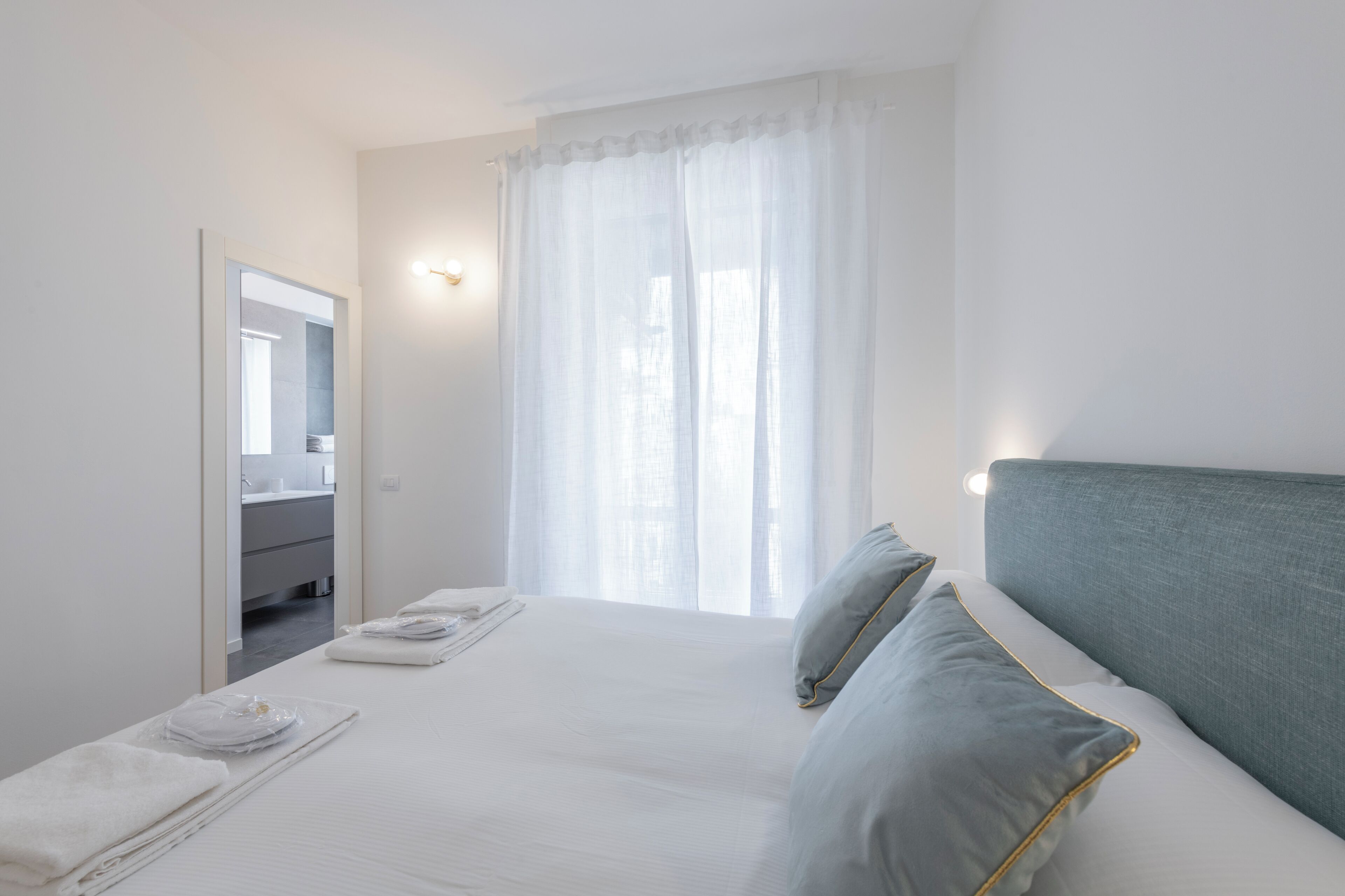 Habitación de hotel con una cama, baño a la vista a través de una puerta abierta y una ventana con cortinas translúcidas.