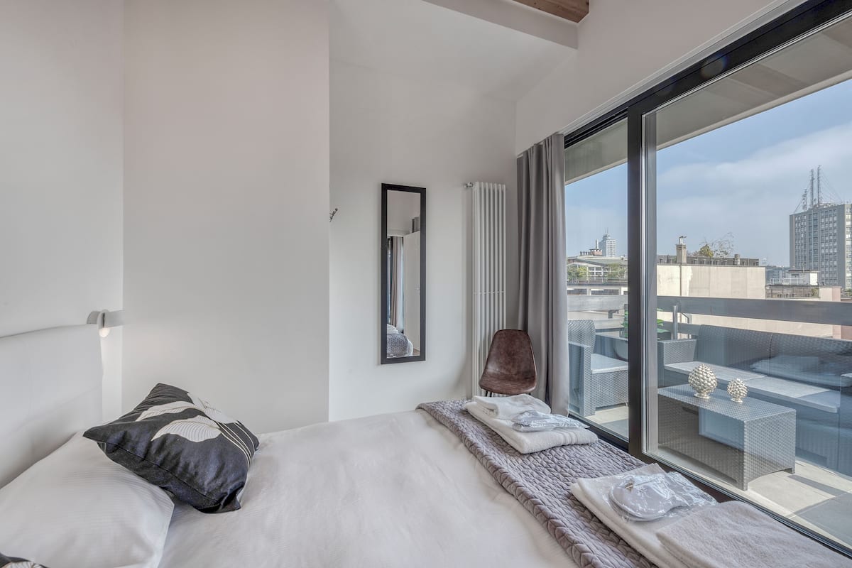 Quarto de hotel moderno com uma cama grande, vista da cidade através de janelas do chão ao teto e uma varanda com mobiliário exterior.