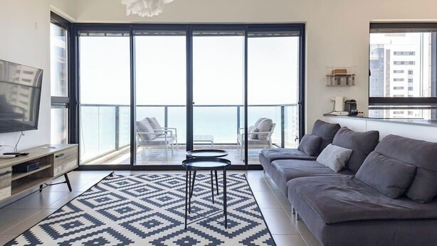 A modern living room with a sofa, a TV, a patterned rug, and a balcony with chairs. - Luxury Apt Tower | Sea View Bat Yam | Mamad (Bat Yam)