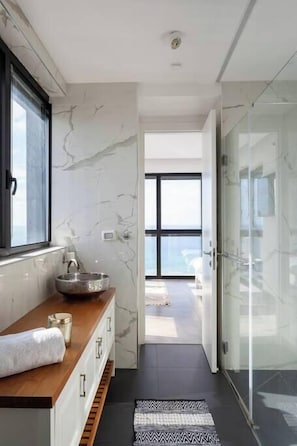 A modern bathroom with a marble wall, a wooden vanity, a glass shower enclosure, and a view of the sky.