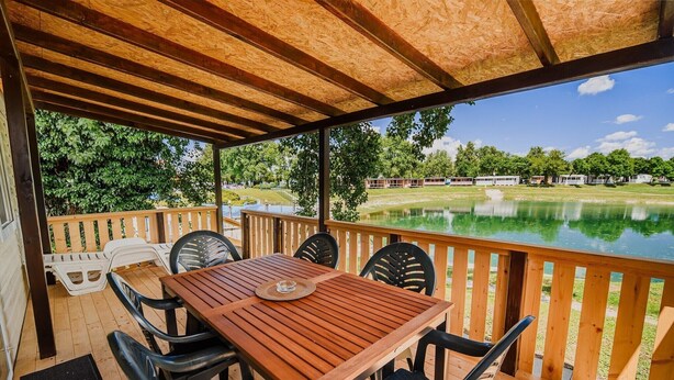 Een overdekt buitengebied met een houten tafel en stoelen, met uitzicht op een meer en een rij bungalows.