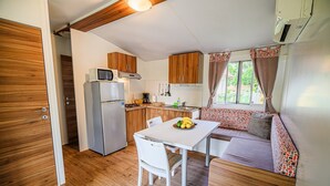 Cocina compacta con armarios de madera, microondas y refrigerador. Hay una mesa de comedor con sillas y un pequeño rincón de estar con un cojín estampado.