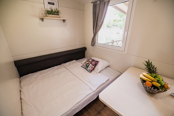 Una cama bien hecha con una almohada y una revista, una mesita con una cesta de frutas, y una ventana con cortinas.