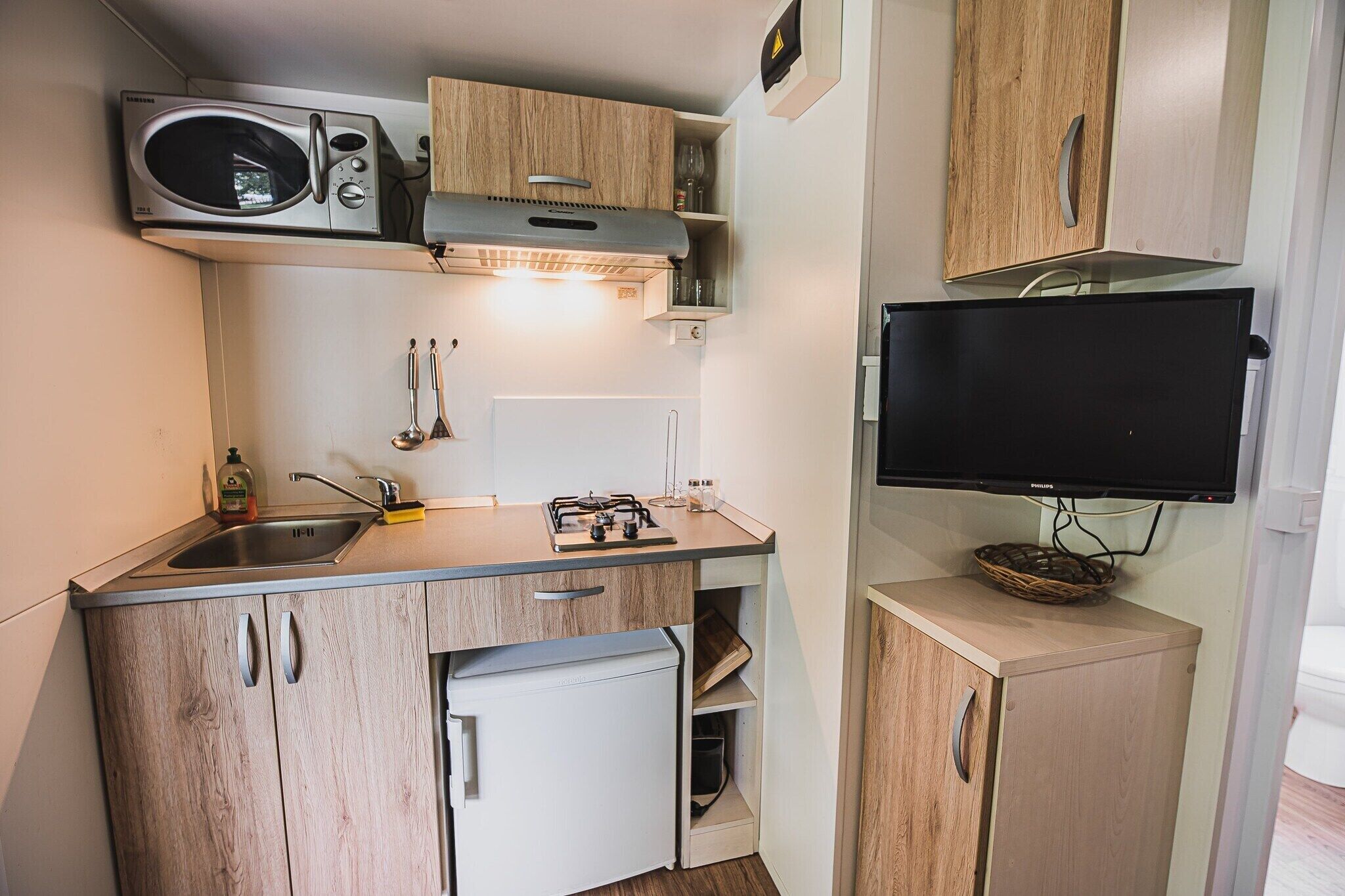 Een kleine keuken met een magnetron, fornuis, spoelbak en een televisie aan de wand.