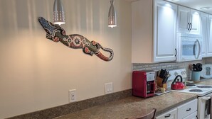 Una cocina con encimera de granito, dos lámparas colgantes, un adorno en la pared, armarios blancos y un hervidor rojo.
