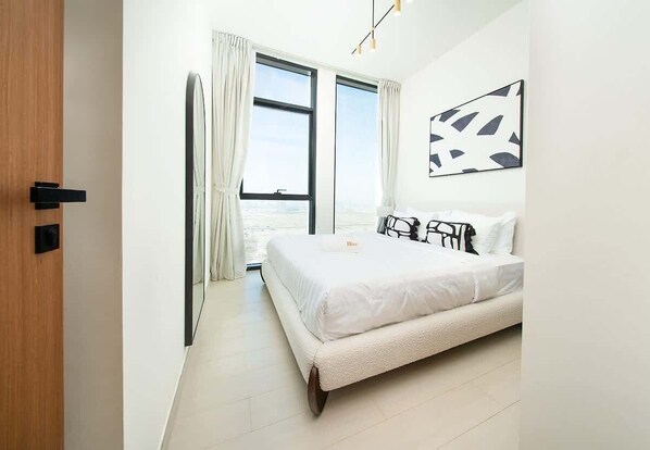 A modern bedroom with a large bed, a view through the window, and a door on the left.