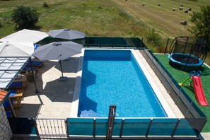 Une piscine rectangulaire aux parois bleues, entourée d’une terrasse verdoyante. On y trouve des parasols et des chaises longues, ainsi qu’un trampoline avec un toboggan situé à droite.