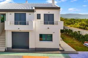 A modern two-story house with a garage, a balcony, and a view of a green landscape.