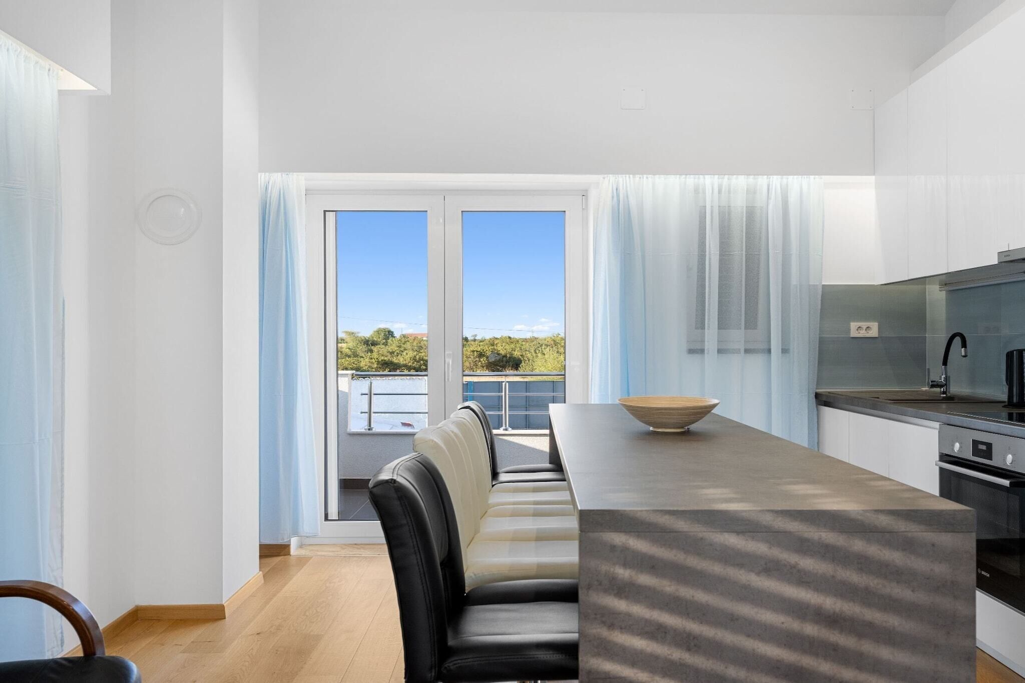 A modern kitchen with a built-in oven, a dining area with a long table and chairs, and a large window with curtains.