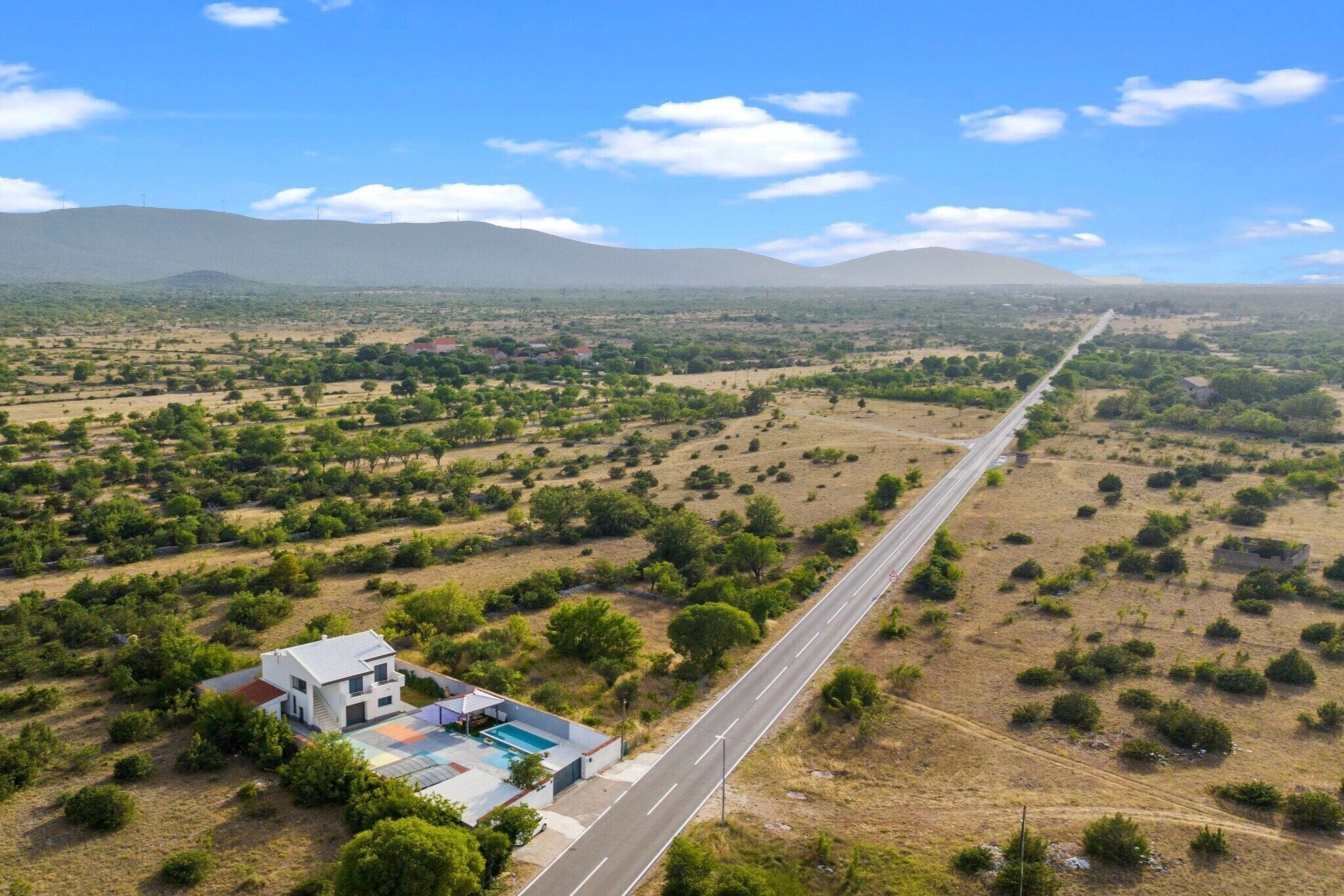 Un edificio moderno de dos pisos con piscina, situado junto a una carretera larga y recta en una zona rural con poca vegetación y montañas al lejano.