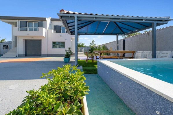 A modern pool area with a covered patio, a building with a garage, and a well-maintained lawn.