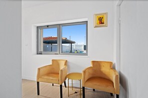 Quarto com uma grande janela, duas poltronas, uma mesa redonda pequena e um quadro na parede.