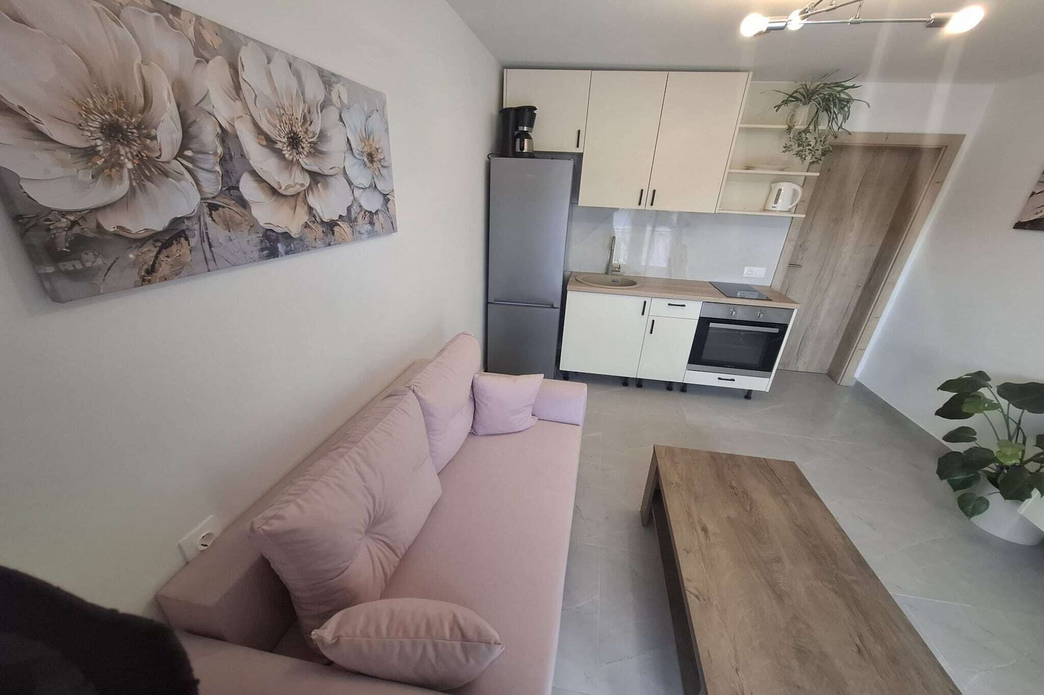 A modern kitchen with white cabinets, a refrigerator, and a microwave. There is a wooden dining table and a beige sofa with cushions.