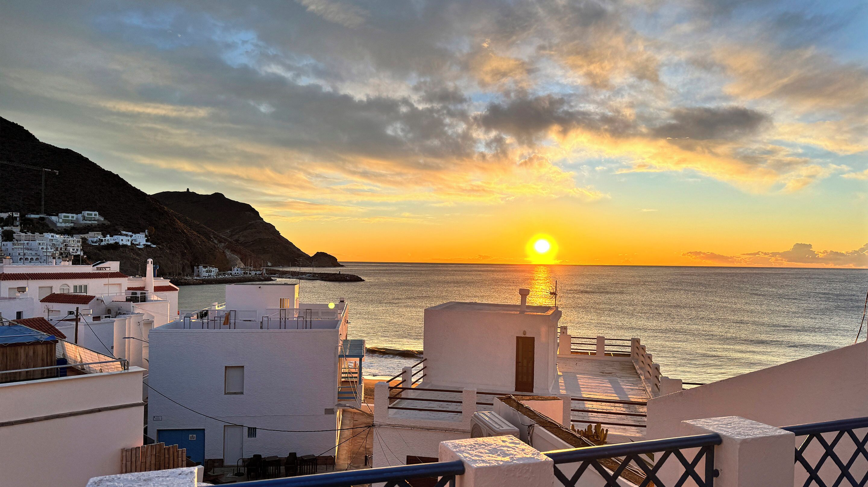 Une vue sur le coucher de soleil en bord de mer, avec des bâtiments au premier plan et une chaîne de montagnes en arrière-plan.