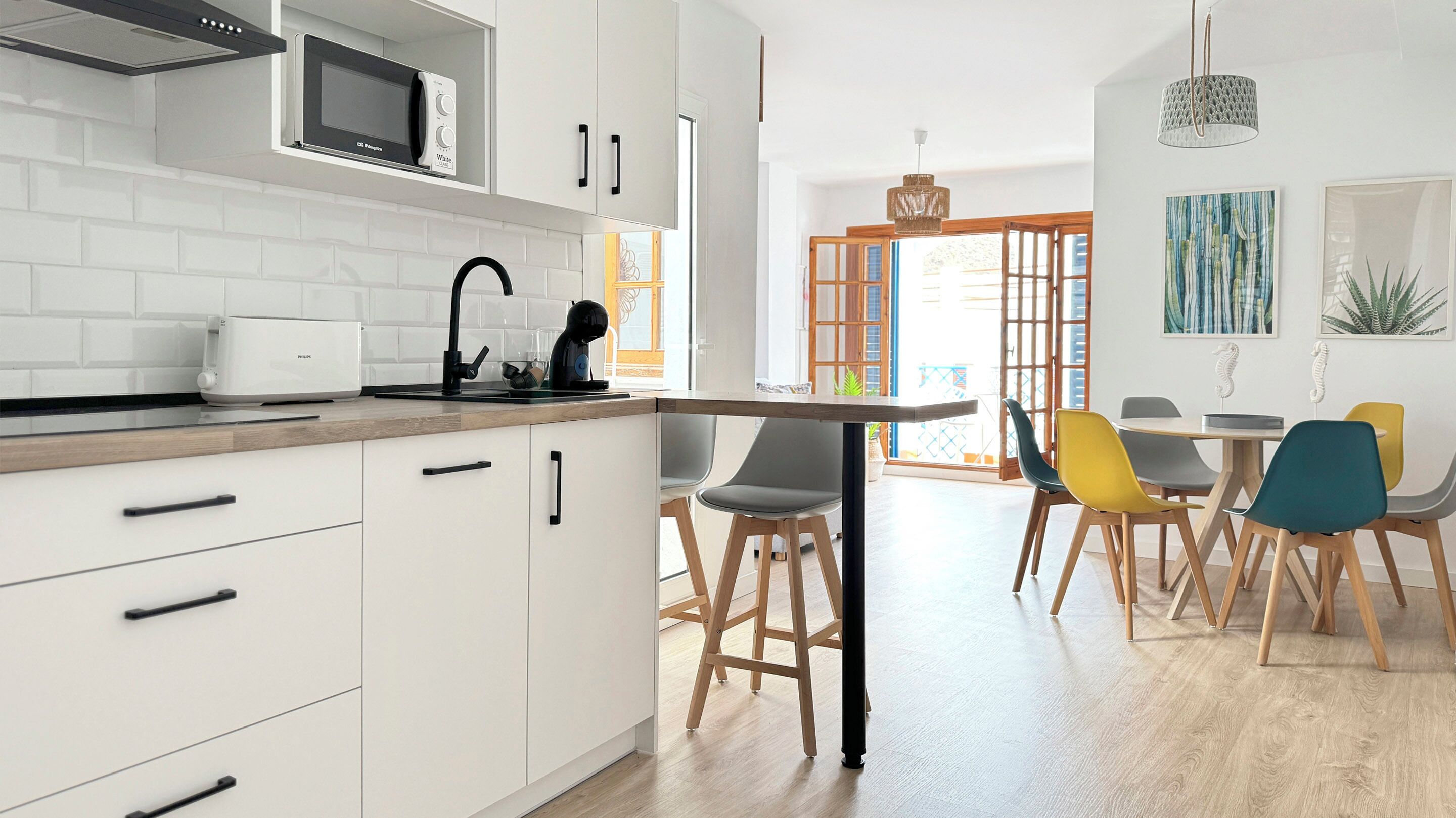 Una cocina moderna con armarios blancos, un microondas y una cafetera. Hay un comedor con una mesa y sillas de colores.