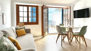 Una sala de estar con sofá, una mesa de comedor con sillas, un televisor montado en la pared y un ventanal con contraventanas.