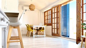 Un coin repas avec une table blanche et des chaises jaunes, une échelle en bois et une grande fenêtre avec des volets en bois.