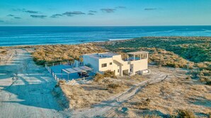 Un bâtiment moderne doté d’un toit plat et de panneaux solaires, situé sur un terrain sablonneux à la végétation clairsemée, et offrant une vue sur une mer calme.