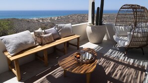 Un balcon avec un banc en bois, des coussins, une chaise en osier, une table ronde et un grand cactus en pot.