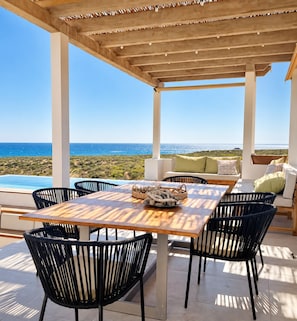 Een overdekt terras met uitzicht op zee, een zwembad en een tafel met stoelen.