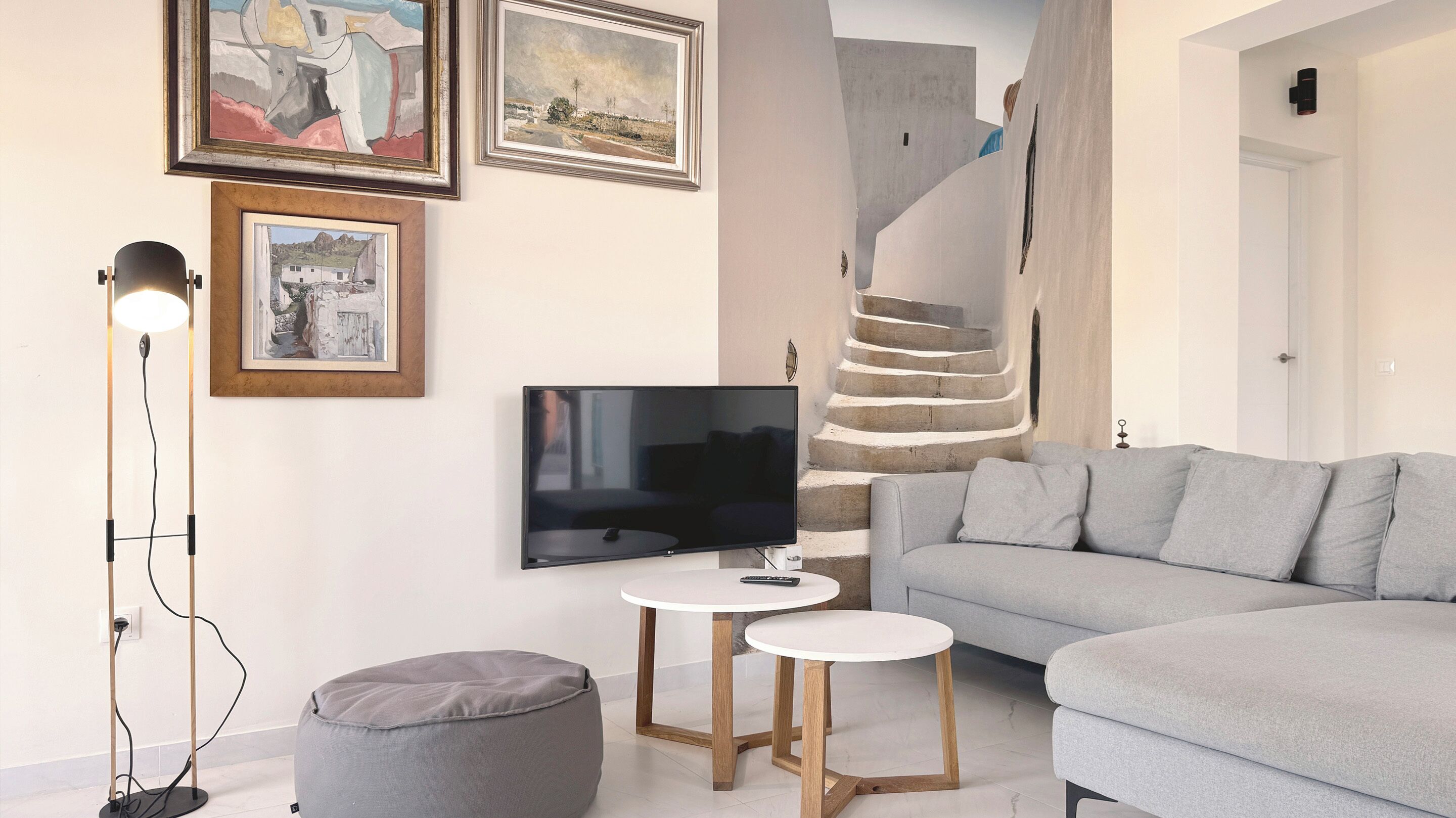 A modern living room with a grey sofa, a flat-screen TV, a floor lamp, and a staircase.