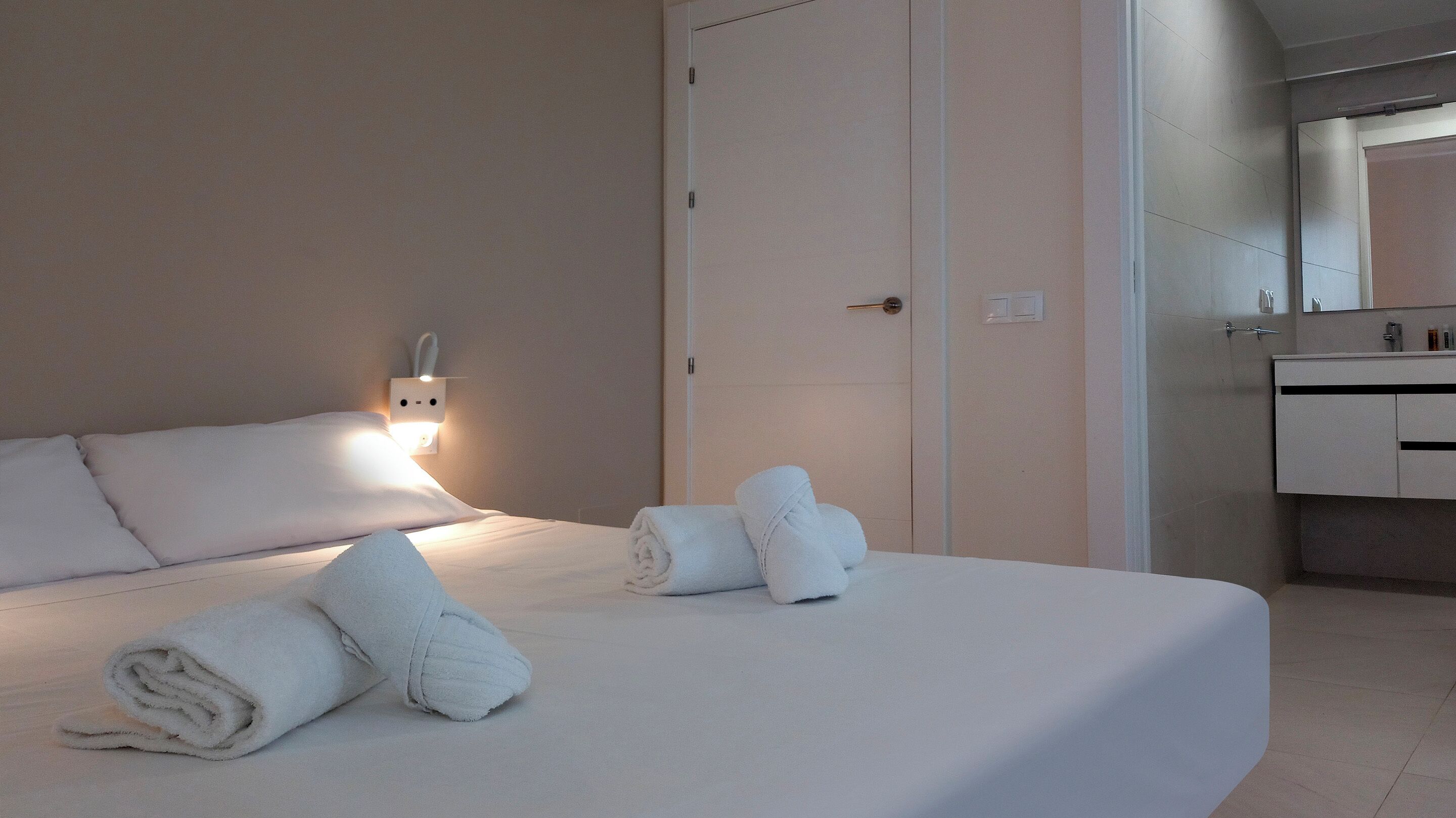 A neatly made bed with rolled towels, a bedside wall socket, and a bathroom with a mirror and sink.