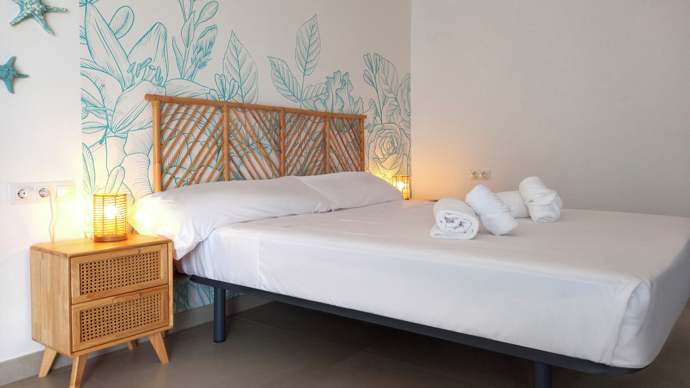 Una cama bien hecha con sábanas blancas, cabecera de madera y mesita de noche con lámpara.