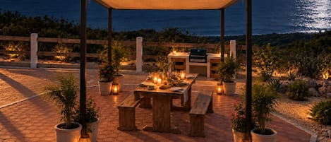 An outdoor dining area with a wooden table and benches, surrounded by potted plants and candles.