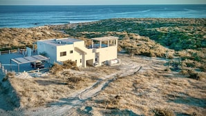 Un edificio moderno a due piani con tetto piatto e un'area esterna coperta, immerso in un terreno collinare e arido con vegetazione rada.