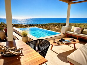 A poolside area with a view of the sea, a wooden table, a black chair, a sofa, and a book titled 'The Mediterranean'.