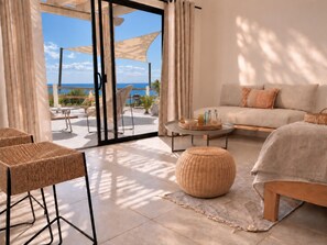 A room with a sofa, ottoman, wicker chairs, a table with drinks, and a view of the sea through large glass doors. - Spacious Serenity Suite · Sea Views · Infinity Pool & Jacuzzi · Walk to Beach (Vinoramas Diamante)