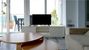 A modern living room with a flat-screen TV, a round wooden table, a beige sofa, and a balcony with a view of the ocean. - Apartamentos Villa Azul (San José)