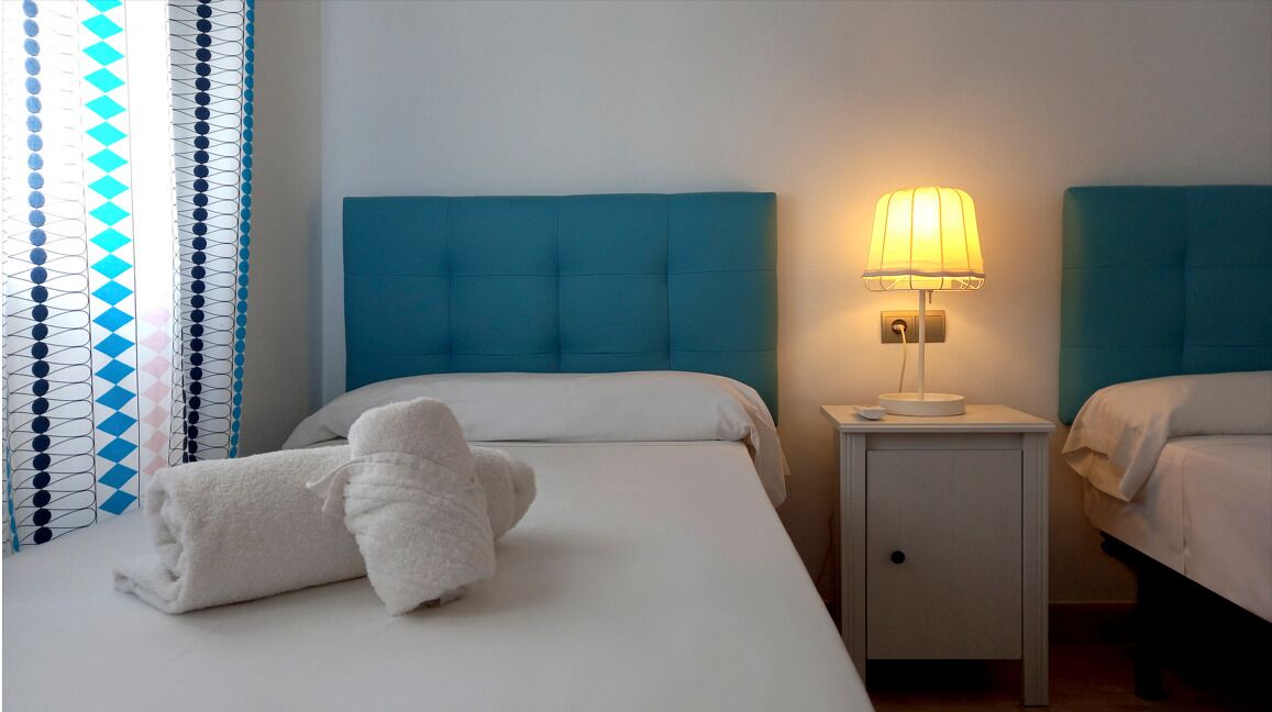 A hotel room with two beds, a nightstand with a lamp, and a rolled-up towel on the bed.