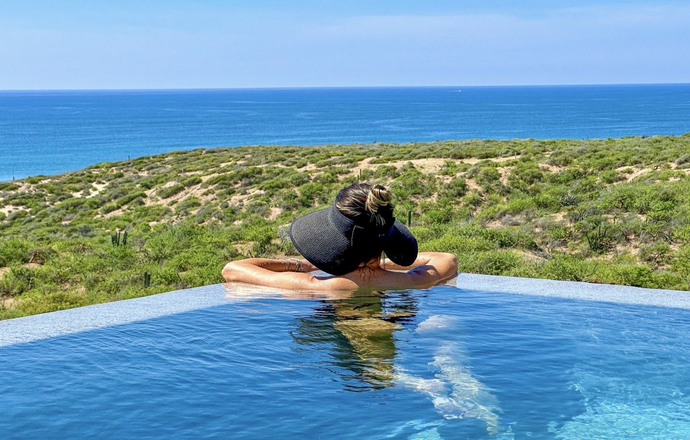 A person sitting in a pool with a view of the ocean and a hilly landscape.