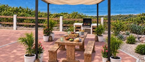 An outdoor patio with a wooden table and benches, a grill, and potted plants.