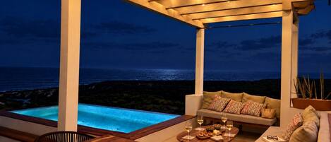 A covered outdoor seating area with a pool, a table with glasses and snacks, and a view of the ocean.