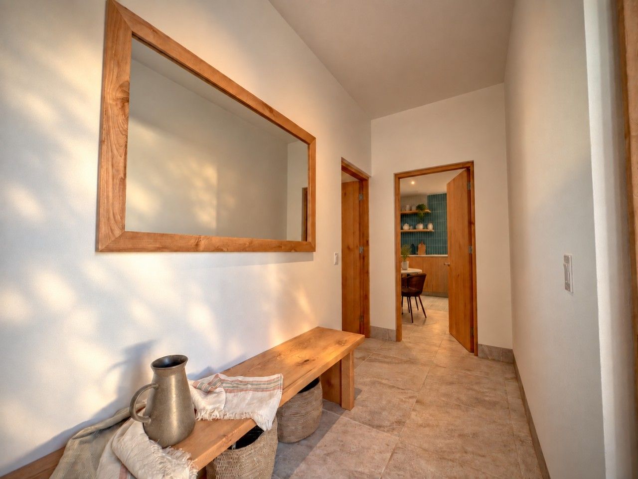 A hallway with a wooden bench, a metal pitcher, and a woven bag. There is a mirror on the wall and a door leading to a room with a blue tiled wall.