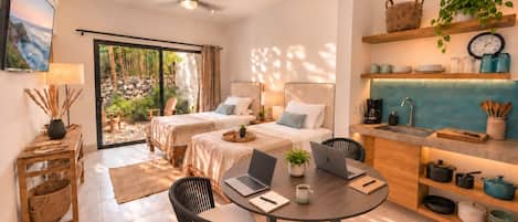 A modern room with two beds, a dining table with laptops, a kitchen area, and a view of the outdoors.