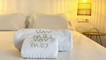 Una cama bien tendida con ropa de cama y toallas blancas, una lámpara de noche y una mesita con una silla.
