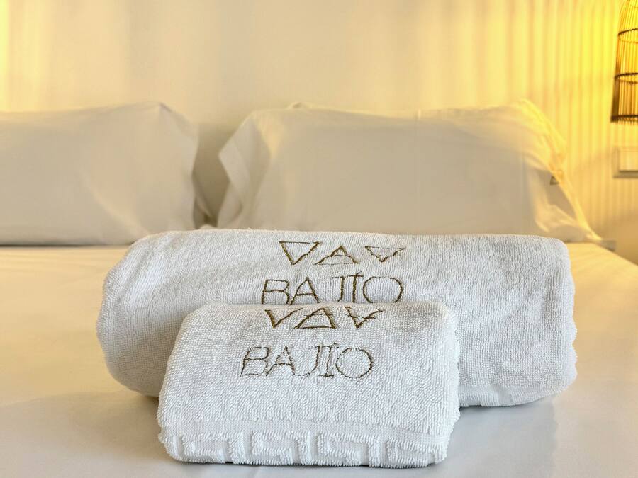 Una cama bien tendida con ropa de cama y toallas blancas, una lámpara de noche y una mesita con una silla.