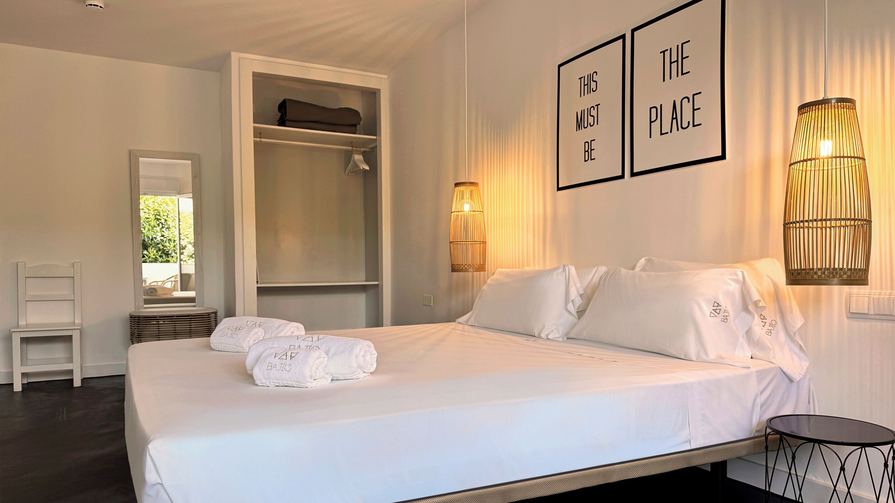 A neatly made bed with white linens and towels, a bedside table, a chair, a wardrobe, and two wall-mounted lamps.