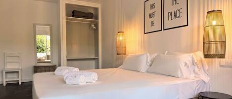 Una cama bien hecha con sábanas y toallas blancas, una mesita de noche, una silla, un armario y dos lámparas fijadas a la pared.