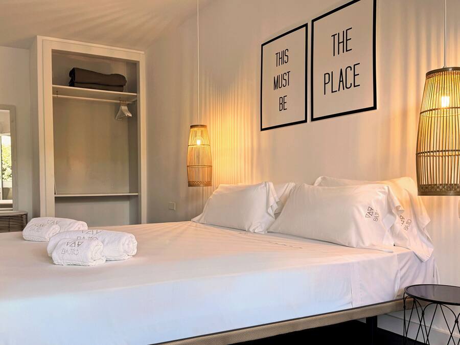 A neatly made bed with white linens and towels, a bedside table, a chair, a wardrobe, and two wall-mounted lamps.