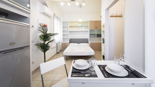 A compact hotel room with a bed, bedside tables, a dining area with a table set for two, and a kitchenette. - Sarpi Flat (Firenze)