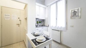A compact kitchen with white cabinets, a dining table set with plates and glasses, a radiator, and a window with curtains. - Sarpi Flat (Firenze)