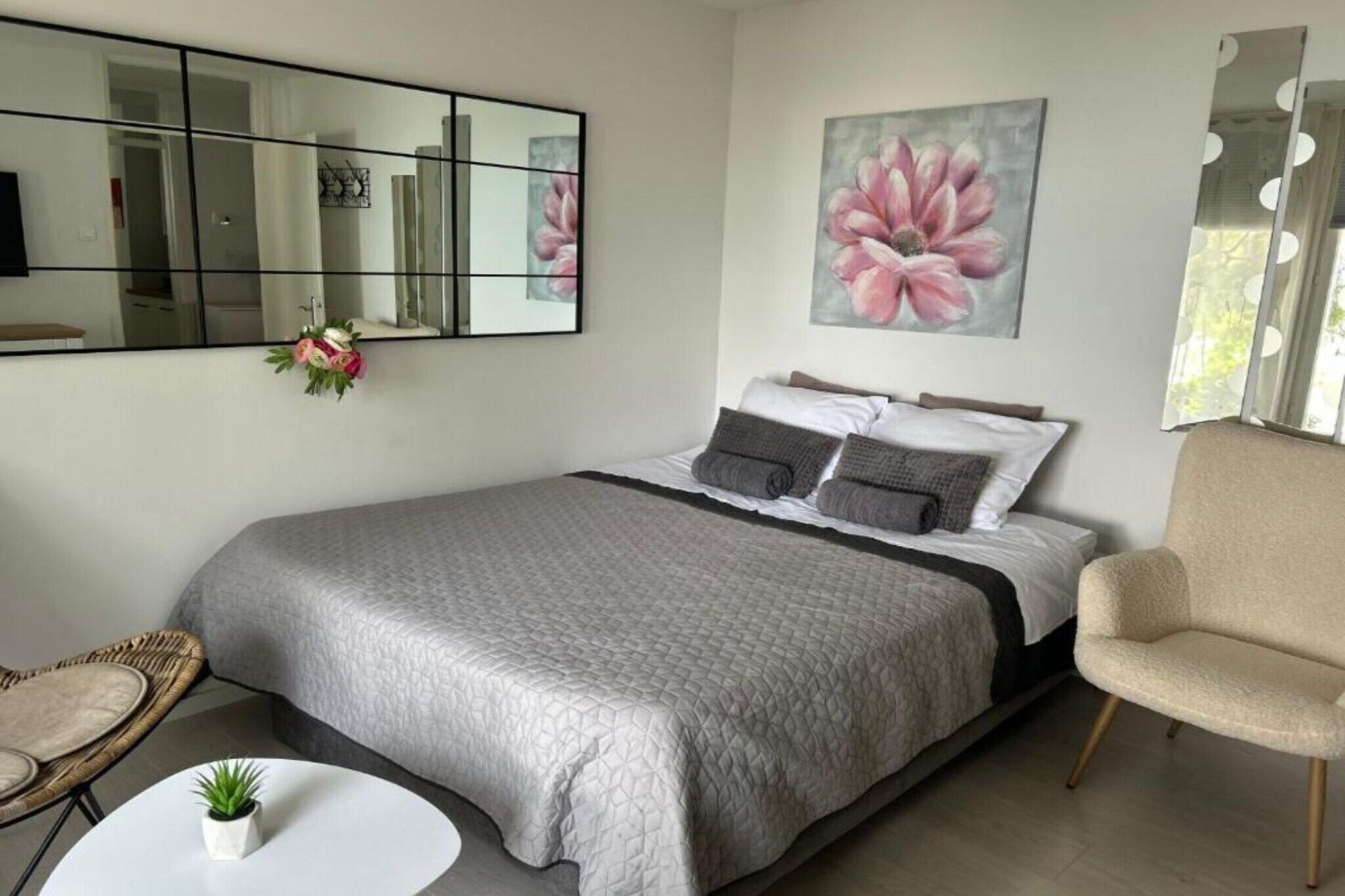 Une chambre à coucher avec un lit, une chaise, une petite table avec une plante, un miroir et un tableau accroché au mur.