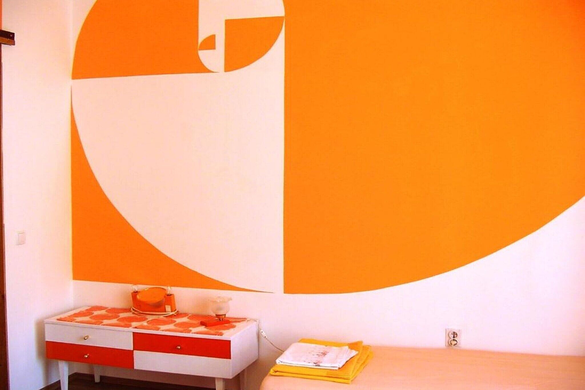 Et værelse med et stort, orange og hvidt abstrakt vægdesign, et lille skrivebord med skuffer og en seng med et gult tæppe.